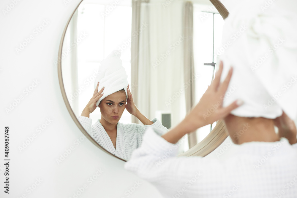 Obraz premium Young woman in bathrobe looking in bathroom mirror. Pure beauty. Attractive woman touching her face and smiling while looking in the mirror