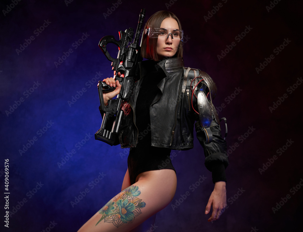 Stylish and beautiful woman with cybernetic hand posing in dark background with spotlight. Dangerous female soldier with short haircut holding rifle.