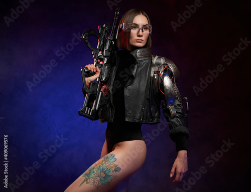 Fototapeta Naklejka Na Ścianę i Meble -  Stylish and beautiful woman with cybernetic hand posing in dark background with spotlight. Dangerous female soldier with short haircut holding rifle.