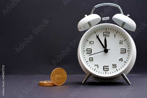 classic white alarm clock stands next to some gold coins and the time shows 5 minutes to 12 photo