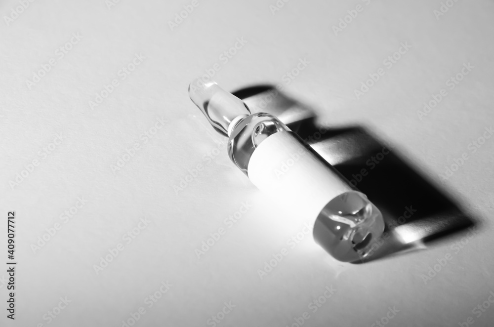 Naklejka premium Black and white photo. Selective focus ampoule with medicine with copy space. The vaccine vial lies on white surface and casts dark shadow. Pandemic concept. Health protection.