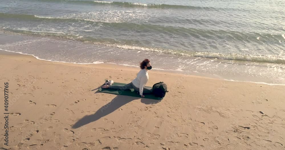 Mujer con mascarilla covid-19 realizando postura yoga en la arena de la playa y dron volando orbital