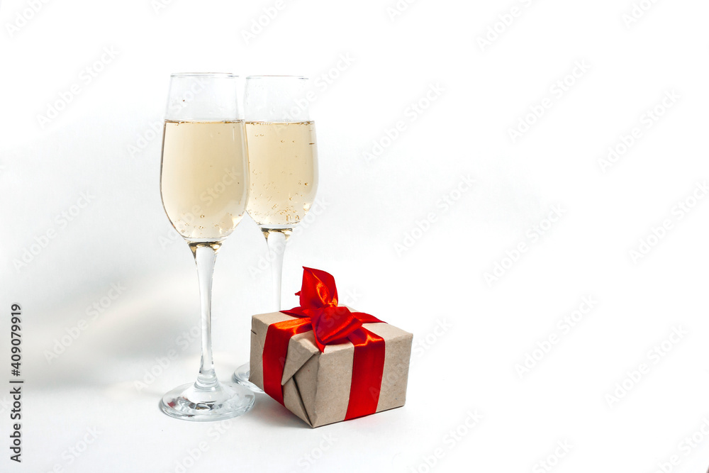 Two glasses of champagne and a gift box tied with a red ribbon on a white minimalist background. copy space Two glasses of champagne and a gift box tied with a red ribbon on a white minimalist background. copy space