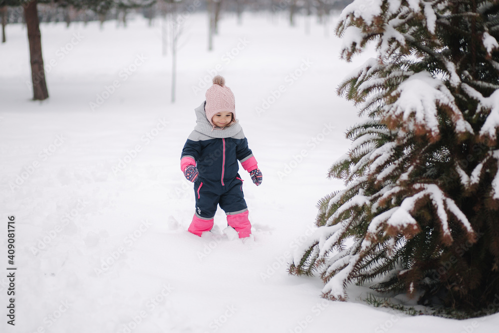 Happy little girl walking in the park on snow. Adorable little girl in a pink hat and overalls walking in the woods on a winter snowy day