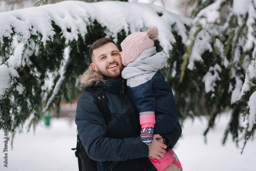 Adorable little daughter with her lovely dad stand by the big snowy tree. Cute little girl laughs and kiss her dad in cheek Adorable little daughter with her lovely dad stand by the big snowy tree. Cute little girl laughs and kiss her dad in cheek