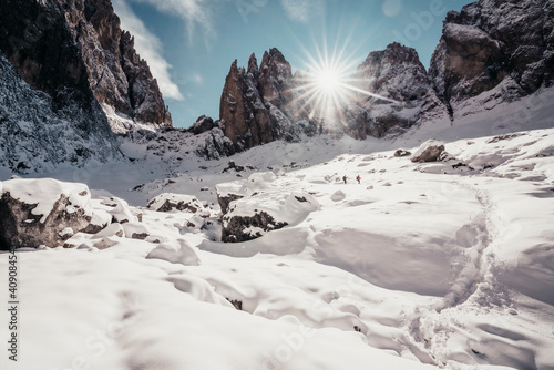 Two hikers descending through rocks on snowy trail in front of Peaks