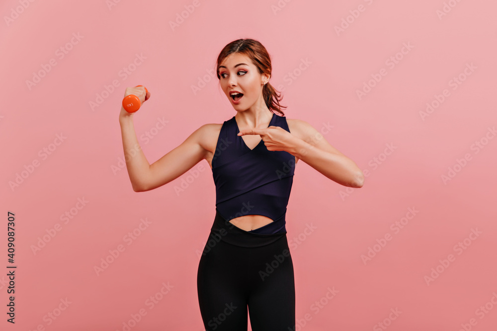 Tanned athlete girl with beautiful makeup in surprise looks at dumbbell and points her finger Tanned athlete girl with beautiful makeup in surprise looks at dumbbell and points her finger