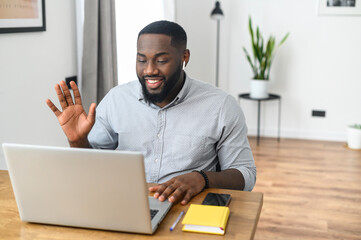 Smiling African American guy wearing airpods speaking close up, using laptop, looking at screen, greeting on video conference, holding a business call working from home. Head shot on light background
