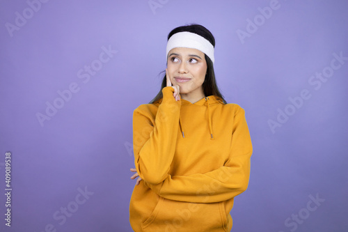 Young latin woman wearing sportswear over purple background with serious face thinking about question with hand on chin, thoughtful about confusing idea © Irene