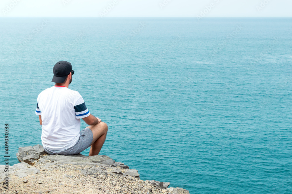 Young man with cap and sunglasses sitting on a cliff in front of the sea. Young man with cap and sunglasses sitting on a cliff in front of the sea.