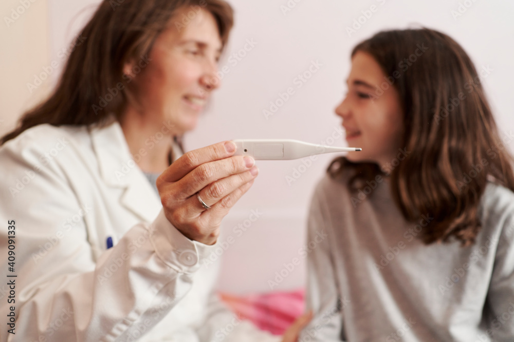 Female doctor teaching the temperature on a thermometer to a girl in her bed. Home doctor concept Female doctor teaching the temperature on a thermometer to a girl in her bed. Home doctor concept