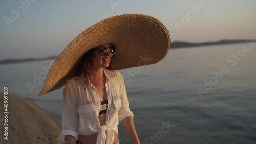 Middle shot of girl in a big hat walking near the sea of Greece and watching the sunset. Young woman wearing a big hat and sunglasses is walking on the coastline of Khalkidhiki island and watching the photo