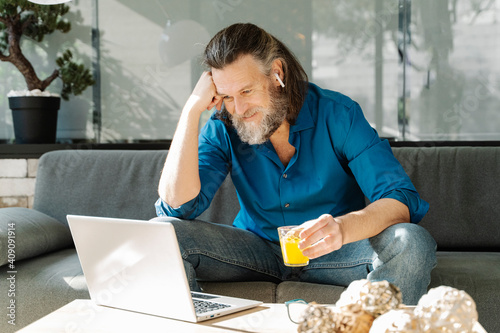 Mature man with a beard drinking orange juice and looking at his lapto photo