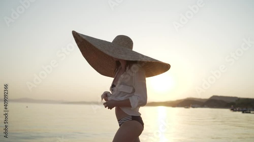 Middle shot of girl in a big hat walking near the sea of Greece and tying up her shirt. Young woman wearing a big hat and sunglasses is walking on the coastline of Khalkidhiki and tying up her shirt photo