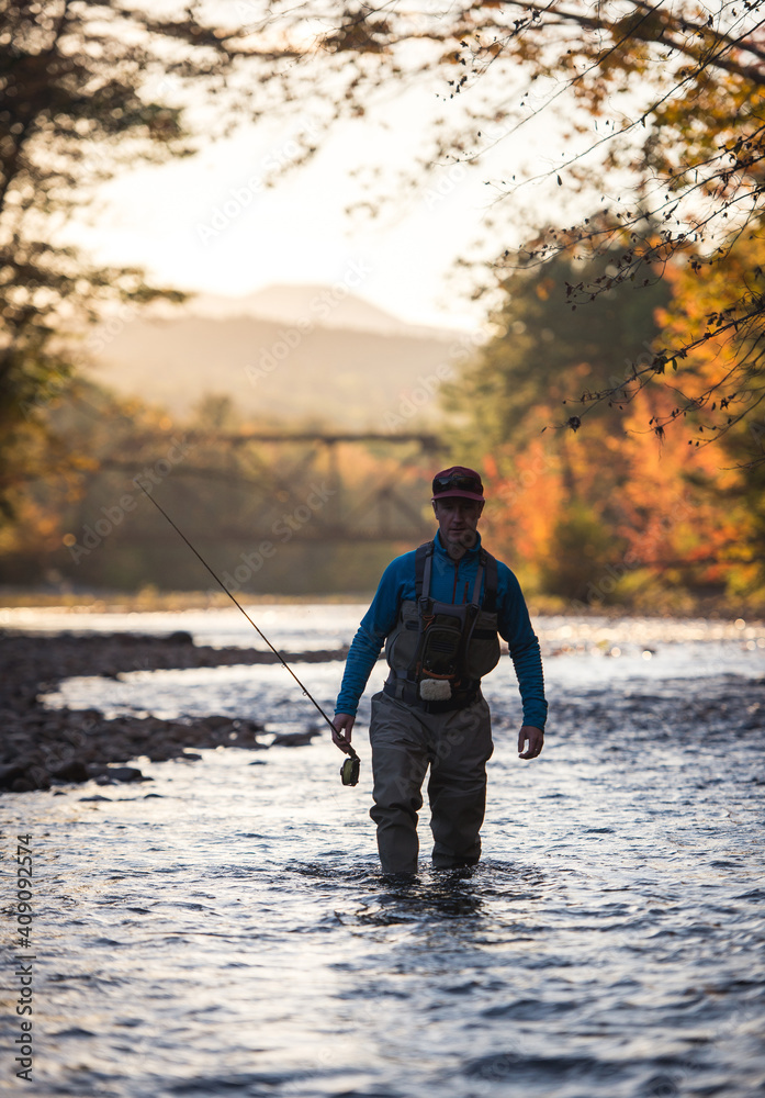 Fly-fisherman wading through river at sunset during foliage season Fly-fisherman wading through river at sunset during foliage season