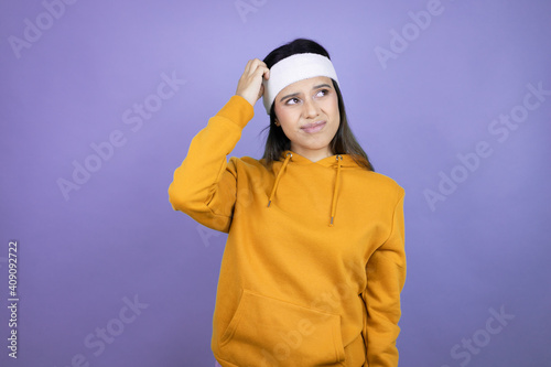 Young latin woman wearing sportswear over purple background confuse and wonder about question. Uncertain with doubt, thinking with hand on head