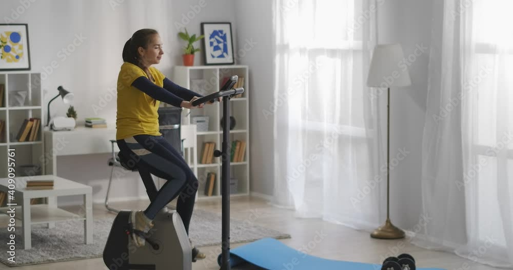 cheerful lady is training alone at home, using exercise bike in living room at daytime, fitness and health, sport activity cheerful lady is training alone at home, using exercise bike in living room at daytime, fitness and health, sport activity