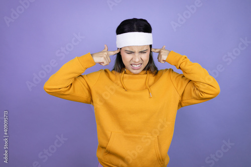Young latin woman wearing sportswear over purple background covering ears with fingers with annoyed expression for the noise of loud music. Deaf concept.