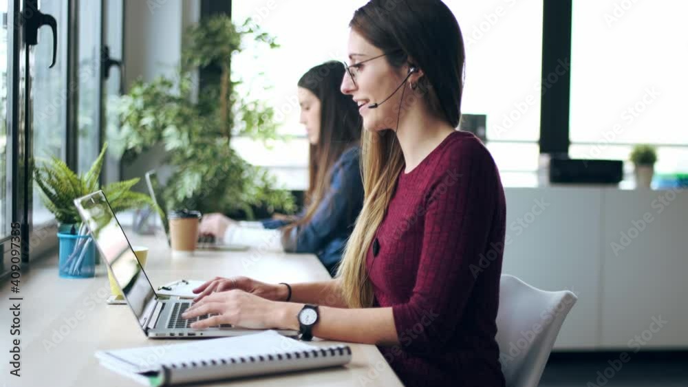 Video of two young business women friends working together with laptop while talking with customers using earphones in the office at home. Video of two young business women friends working together with laptop while talking with customers using earphones in the office at home.