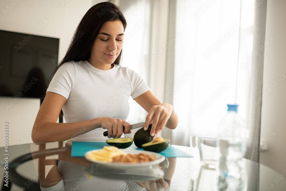 Content female preparing snack of avocado sitting in kitchen Content female preparing snack of avocado sitting in kitchen
