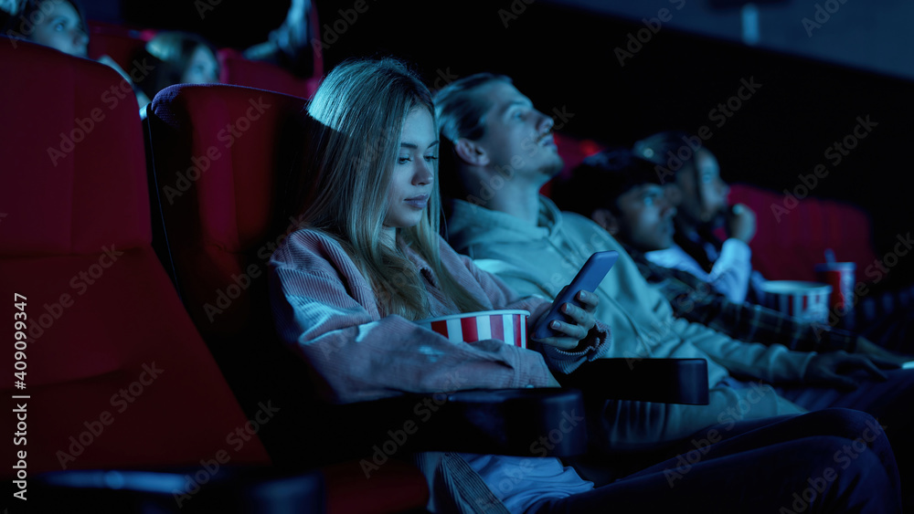 Beautiful young woman using her phone for texting while missing boring movie at the cinema