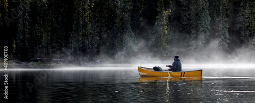 Man paddles canoe on lake with mist and fog on sunny day by forest