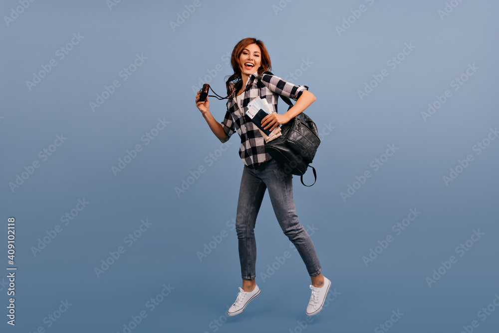 Pretty budget tourist in hurry for tour. Woman with backpack behind her shoulders and in jeans runs on blue background