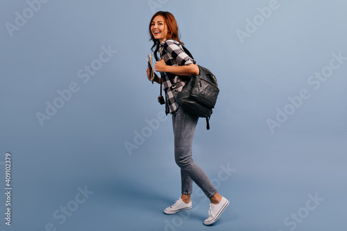 Full-length shot of travel blogger with huge backpack. Girl in converse joyfully moves on isolated blue background