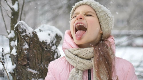 A girl playing with snow in winter, eating snow with tongue and closed eyes outdoors in winter clothes. Happy childhood and active holidays concept