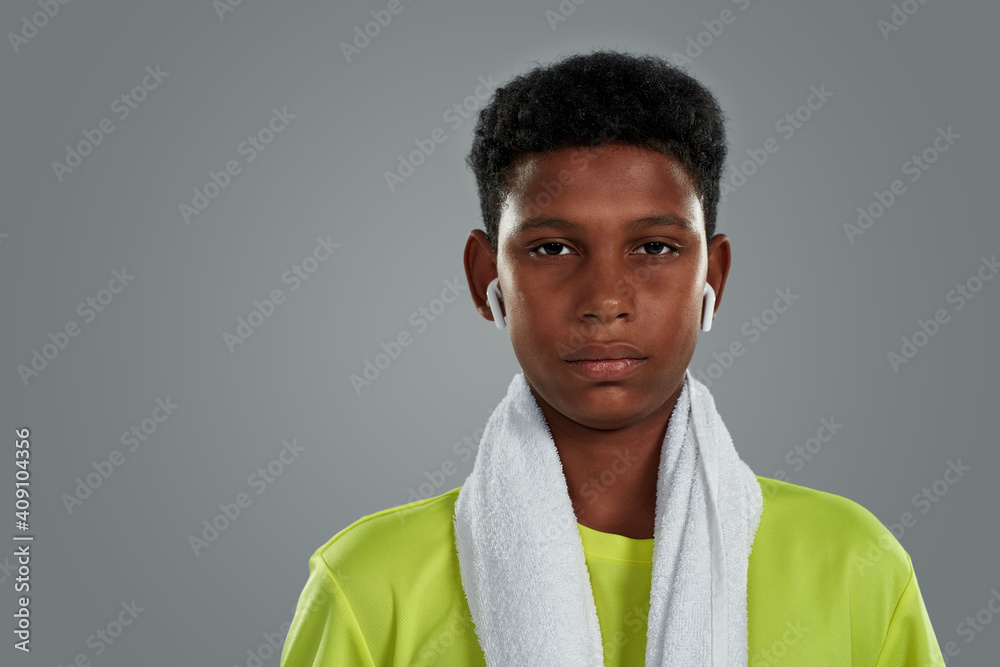 Obraz premium Active lifestyle. Close up portrait of a confident teenage african boy in neon shirt with white towel on shoulders wearing wireless earphones looking at camer