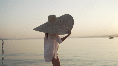 Middle shot of girl in a big hat walking near the sea of Greece and turning her head to watch the sunset. Young woman wearing a big hat and sunglasses is walking on the coastline of Khalkidhiki island photo