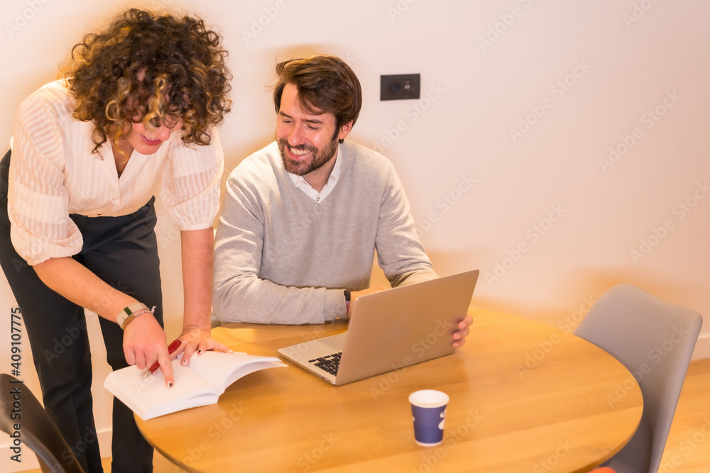 Very good atmosphere in the office between the female boss and the young worker at a table in the office, with a computer and a notebook Very good atmosphere in the office between the female boss and the young worker at a table in the office, with a computer and a notebook