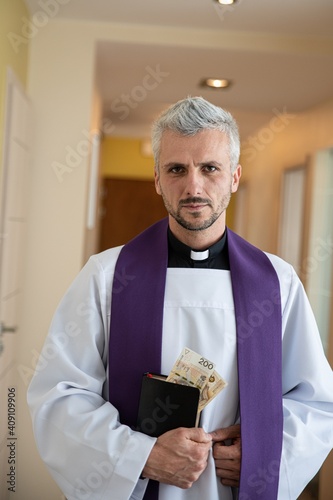 During a pastoral visit, a handsome young priest holds an envelope with money in the Holy Bible. Polish tradition.