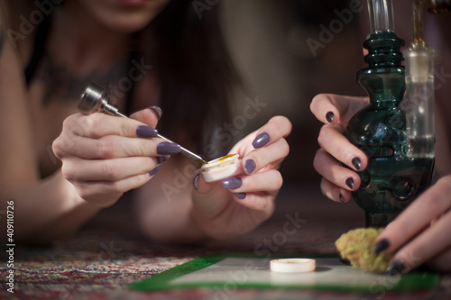Women with nicely manicured fingernails use a titanium dabber tool to scoop concentrated cannabis wax, shatter, oil out of silicone container to smoke in a chilled custom blown glass water pipe design