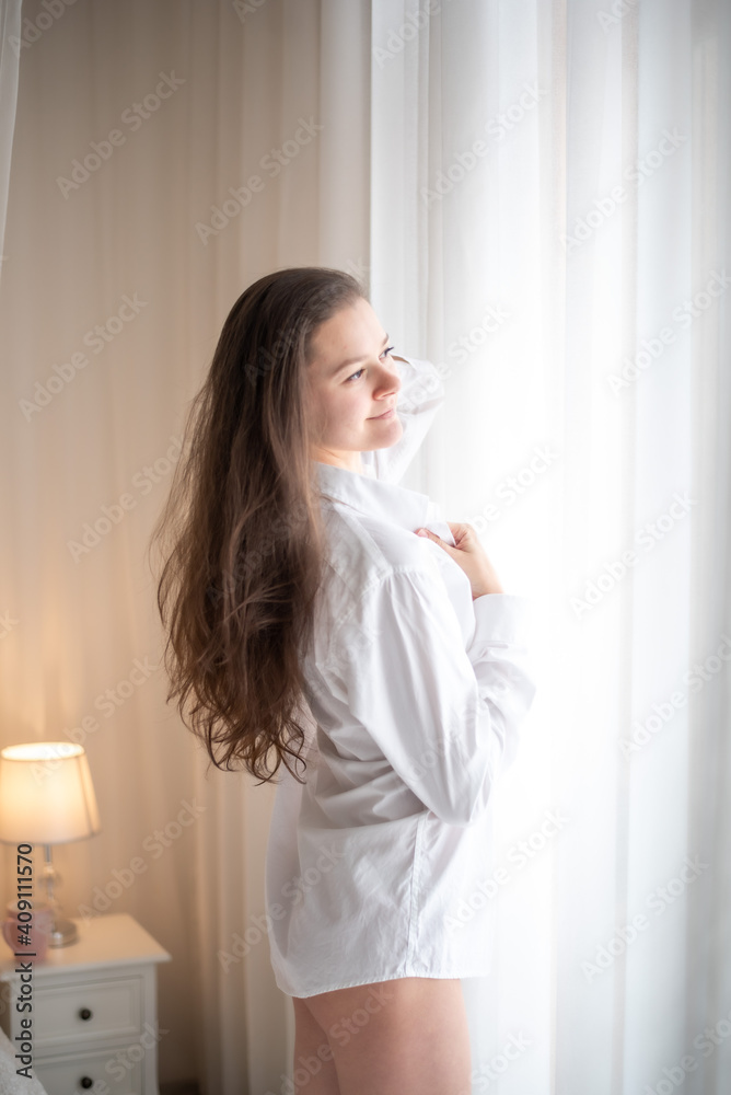 Brunette in mens shirt. Rear view of young woman looking through the window in the morning Brunette in mens shirt. Rear view of young woman looking through the window in the morning
