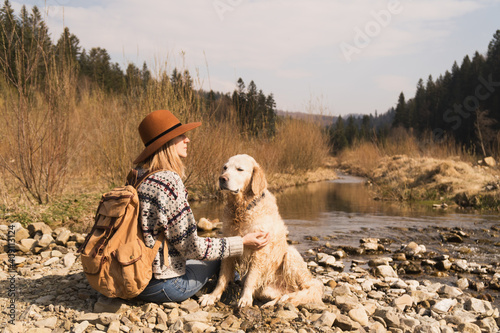 Traveling woman and golden retriever dog in nature. Stylish hipster in brown hat and wool sweater with vintage textile backpack near mountains river and forest.  Travel and wanderlust concept. © prystai