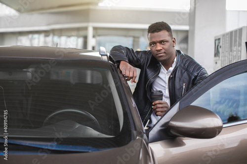 Petrol station, car refueling concept. Positive African man in casual outfit leaning on his car door and looking at camera while standing on blurred background of gas station © sofiko14