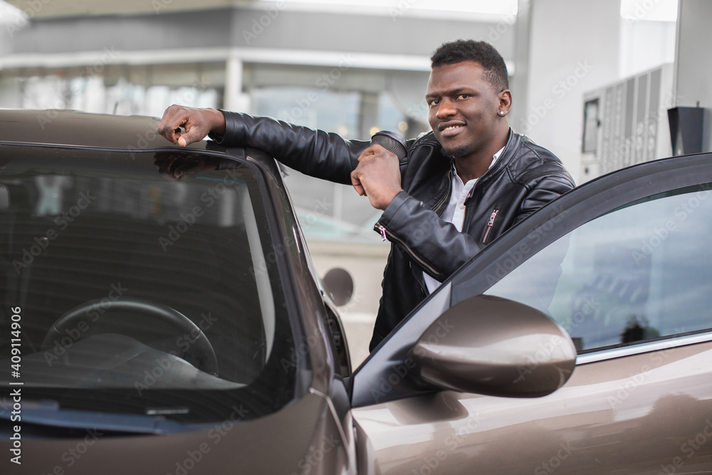 Happy smiling young black man, holding take away coffee cup, and leaning on his car, while posing to camera at petrol station