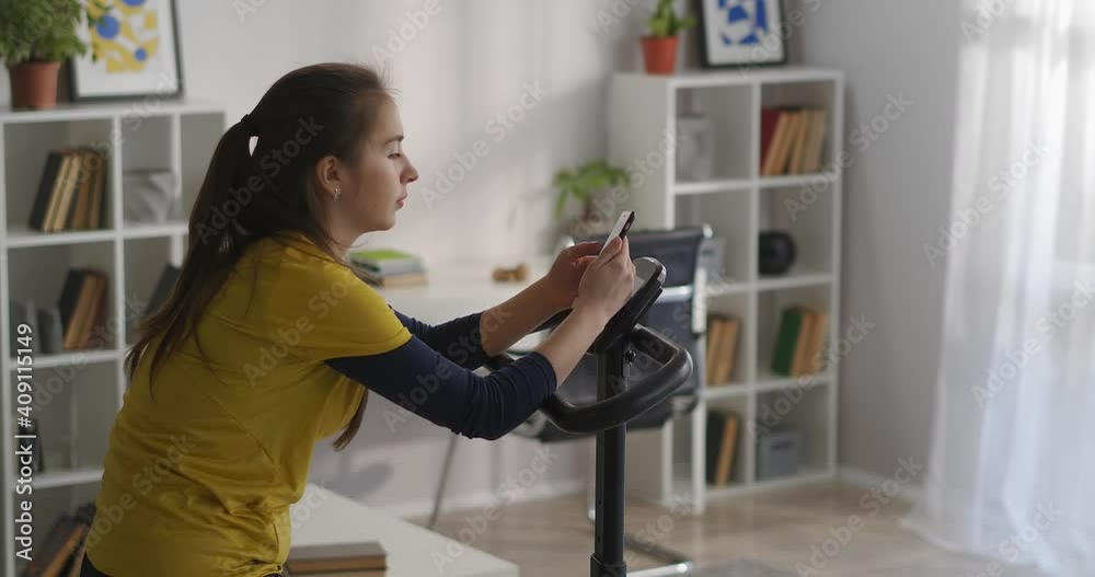 woman with smartphone is sitting on stationary bike at home, viewing social nets and choosing music for workout