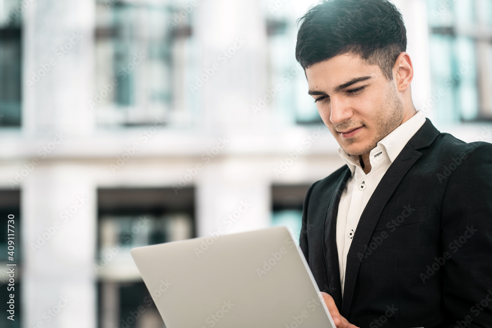 a dark-haired businessman of European appearance stands in a business center in a stylish black suit. holds in his hands works on a laptop computer. copy space. banner for the site. a dark-haired businessman of European appearance stands in a business center in a stylish black suit. holds in his hands works on a laptop computer. copy space. banner for the site.