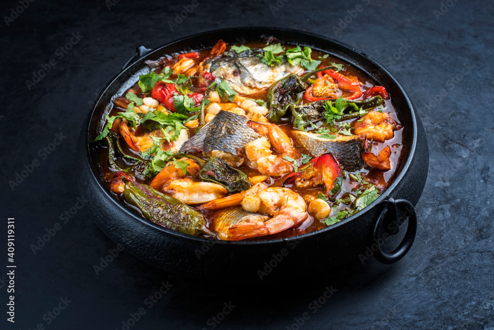 Modern style traditional Mexican seafood pozole soup with fish, king prawns and hominy in a clear sauce served as close-up in design pot Modern style traditional Mexican seafood pozole soup with fish, king prawns and hominy in a clear sauce served as close-up in design pot