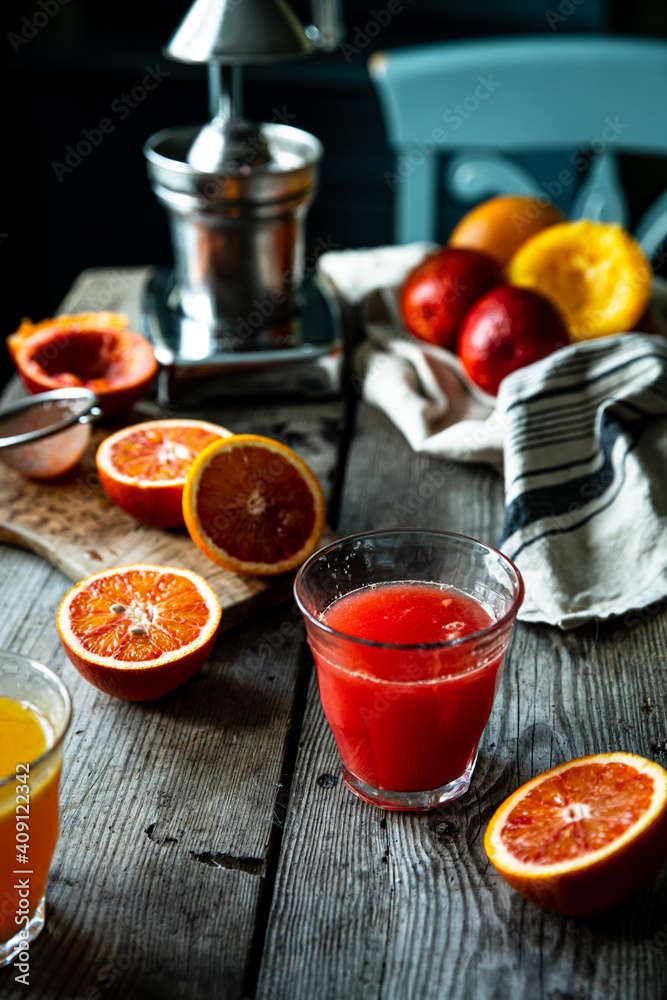 Glass of fresh blood orange juice, orange halves and juicer machine on wooden table with chair on background . Glass of fresh blood orange juice, orange halves and juicer machine on wooden table with chair on background .
