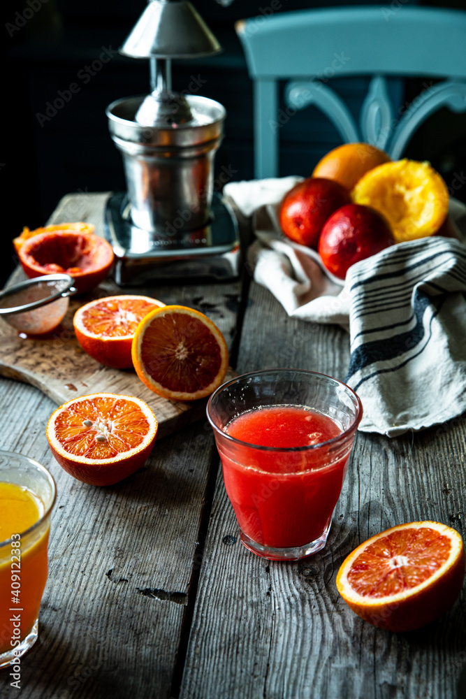 Glass of fresh blood orange juice, orange halves and juicer machine on wooden table with chair on background . Glass of fresh blood orange juice, orange halves and juicer machine on wooden table with chair on background .
