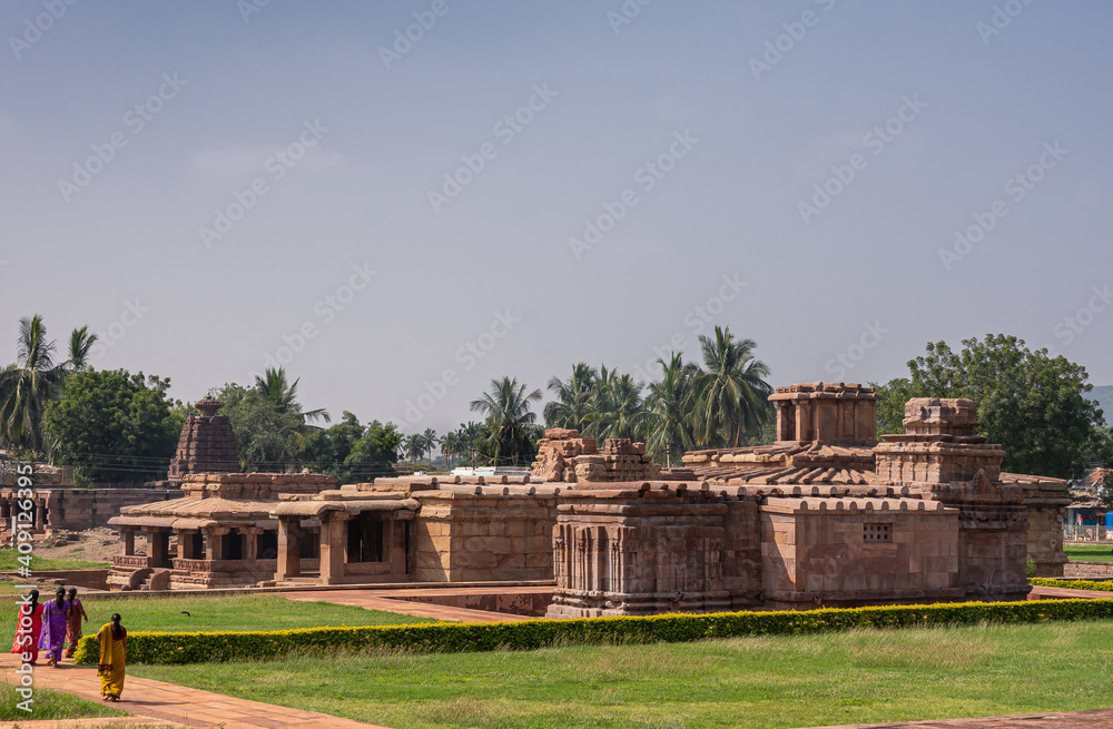 Aihole, Karnataka, India - November 7, 2013: Brown stone Lad Khan and Suryanarayana Temples landscape with green lawns under light blue sky. Saris of women add colors. Green foliage in back.