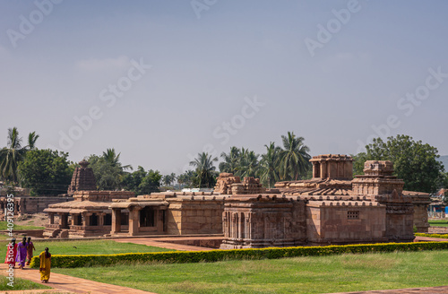 Aihole, Karnataka, India - November 7, 2013: Brown stone Lad Khan and Suryanarayana Temples landscape with green lawns under light blue sky. Saris of women add colors. Green foliage in back. photo