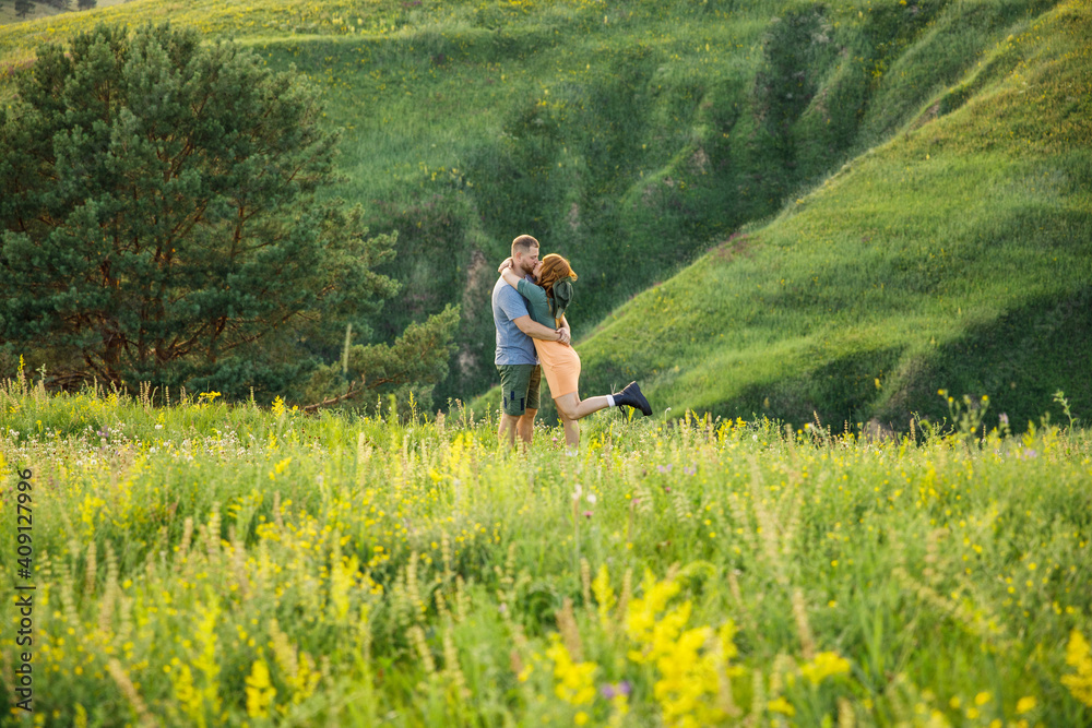 young beautiful couple red-haired girl in a pink dress and green jacket a man in a gray t-shirt and green shorts are having fun in the grass in a field in nature at sunset