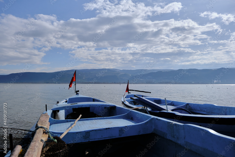 Fototapeta premium Small and blue boats near the coast of Lake of Uluabat in Turkey. Small Turkish flag on the boat with lake and mountain background. Bursa. Golyazi (Apollyon). Bursa, Turkey. 22.01.2021.
