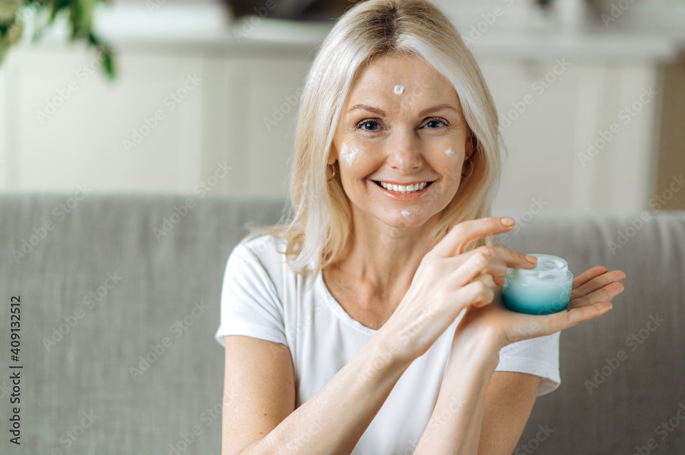 Smiling blonde senior woman apply anti-aging cream to her face. Happy mature woman using cosmetic cream to hide wrinkles on face, moisturizes and nourishes the skin Smiling blonde senior woman apply anti-aging cream to her face. Happy mature woman using cosmetic cream to hide wrinkles on face, moisturizes and nourishes the skin