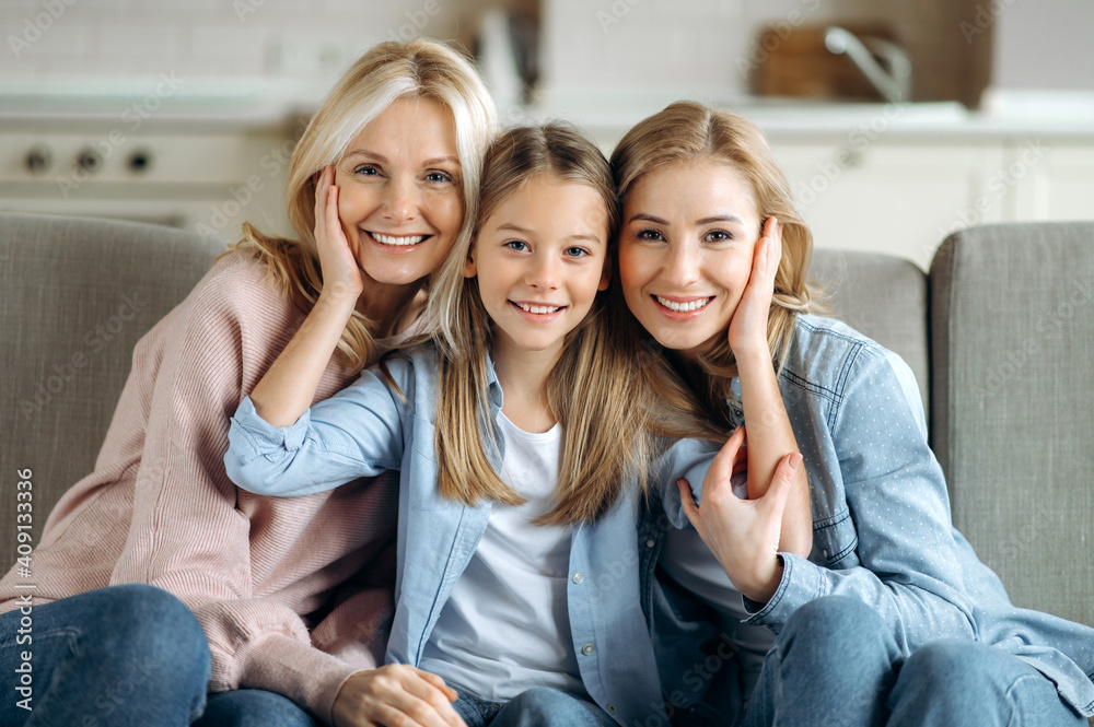 Three female generation. Happy caucasian grandmother, daughter and granddaughter are sitting at home on the couch, hugging, looking at the camera and smiling. Family relationships and values Three female generation. Happy caucasian grandmother, daughter and granddaughter are sitting at home on the couch, hugging, looking at the camera and smiling. Family relationships and values