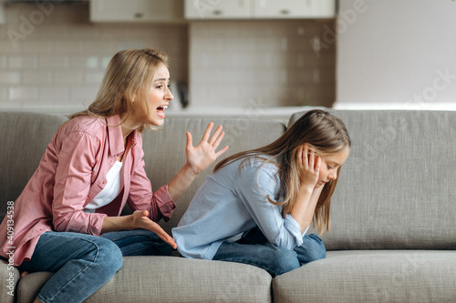 Angry mother scolding little daughter at living room, parent teaches a naughty mischievous child, kid girl feels upset about punishment and deprivation of entertainment photo